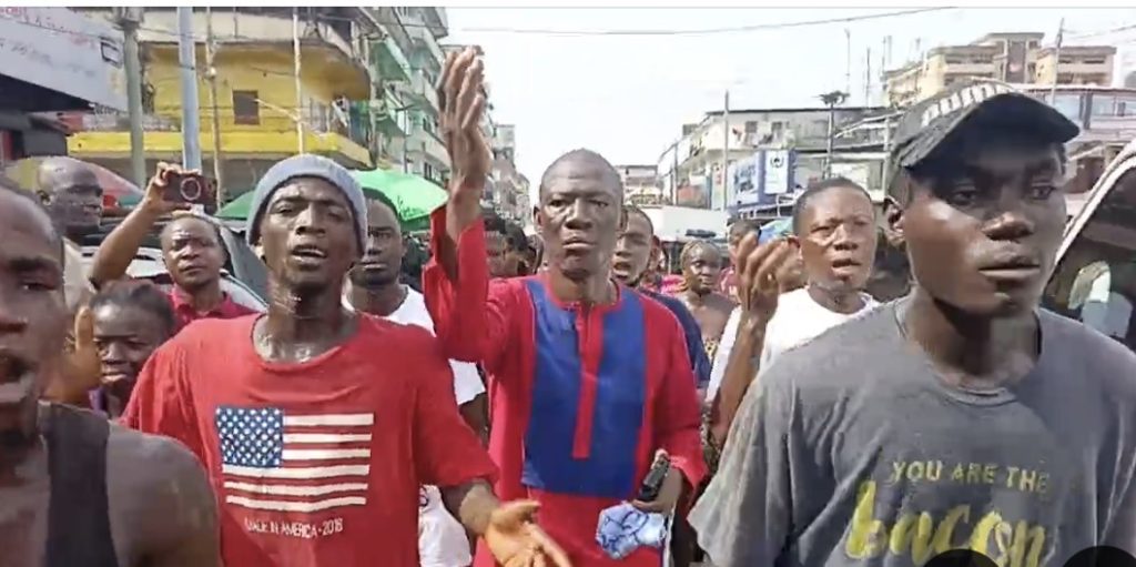 A crowd of men shouting and raising hands in a street rally, with colorful shirts and storefronts in the background.